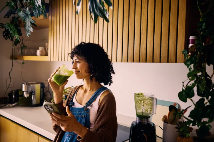 A Young Nutritionist at Home Drinking a Nutritious Smoothie with Fresh Produce to Enhance Metabolism and Vitamin Intake, Planning Her Weekly Meals on Her Smartphone